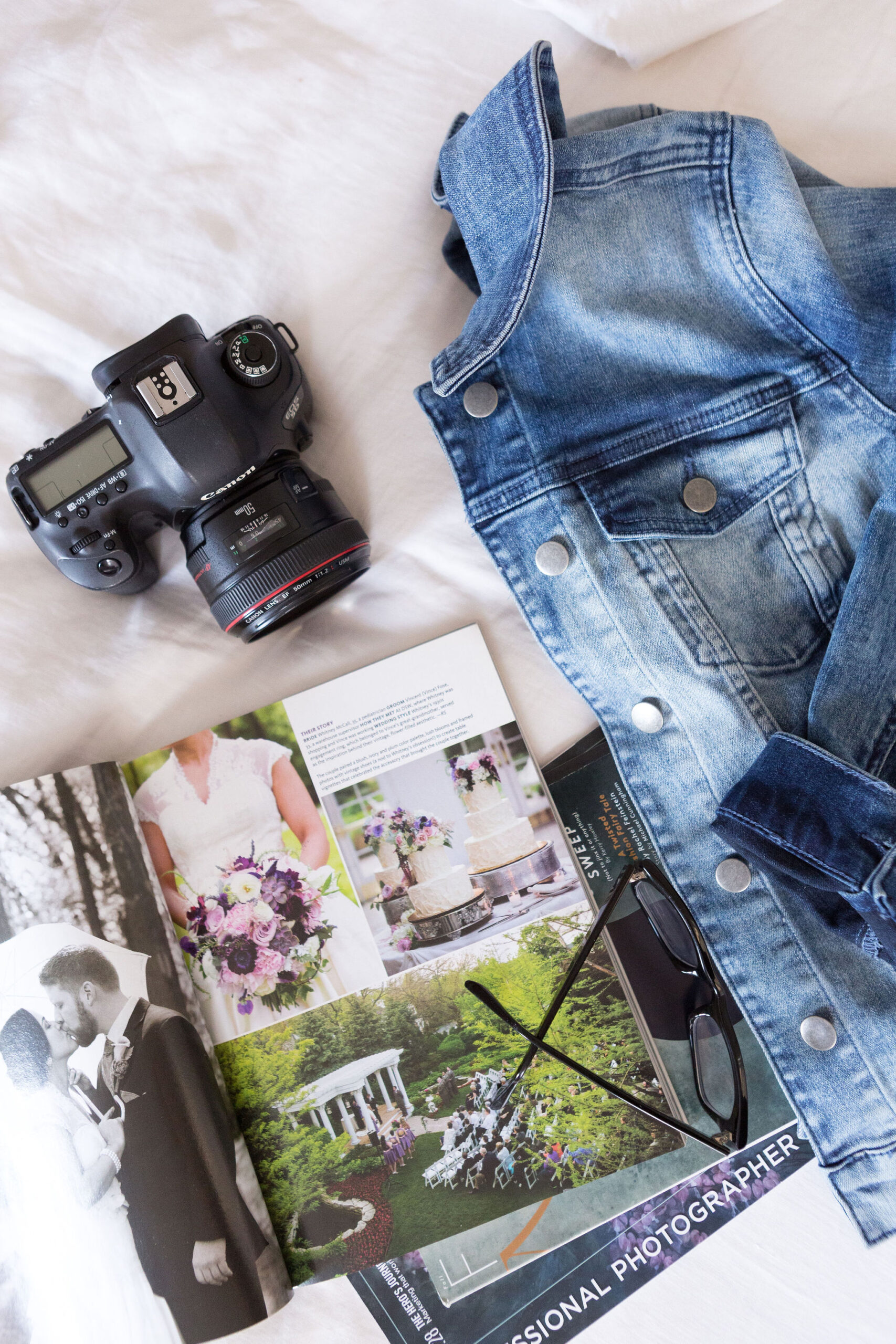Professional camera, wedding magazine and jean jacket laying on a bed.