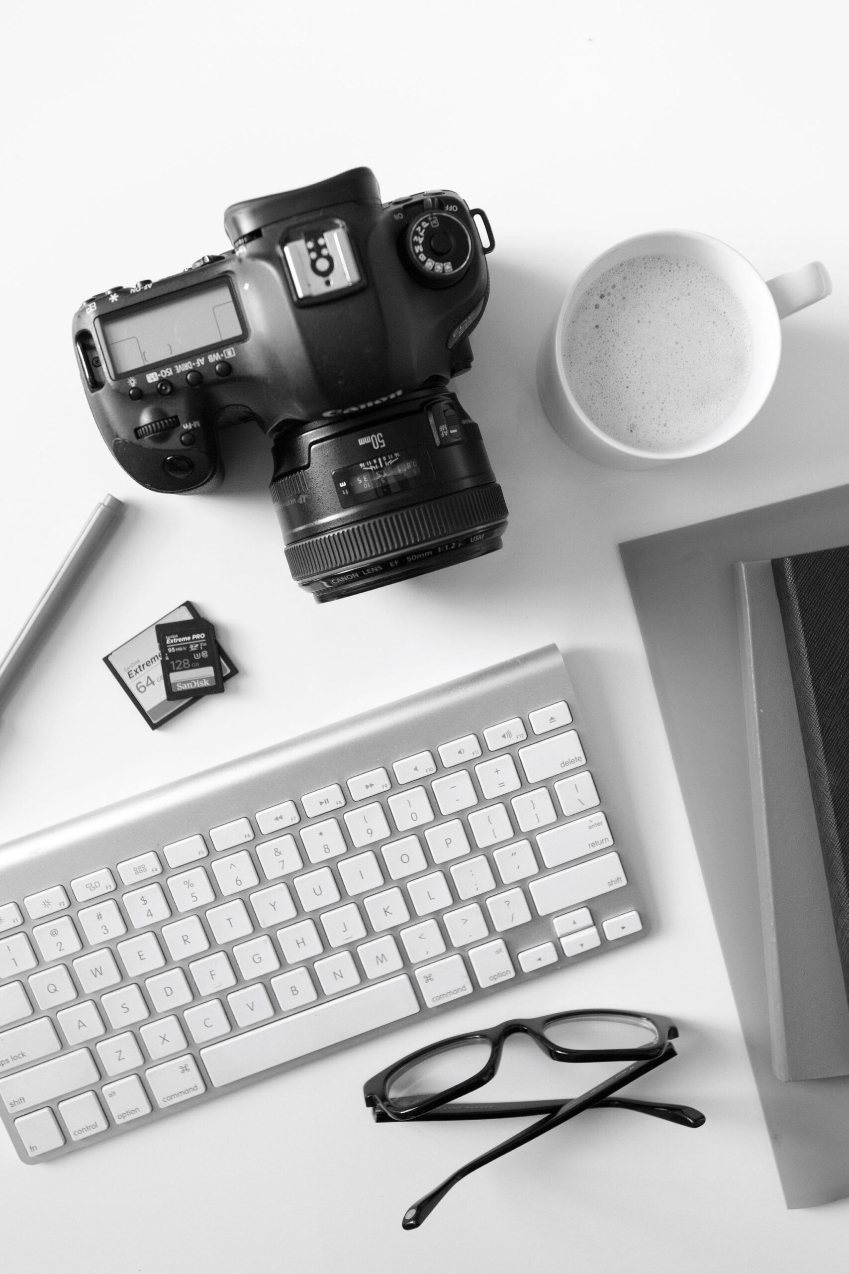 Black and white photo of a professional photographer's camera on a desk with a coffee, keyboard and memory cards.