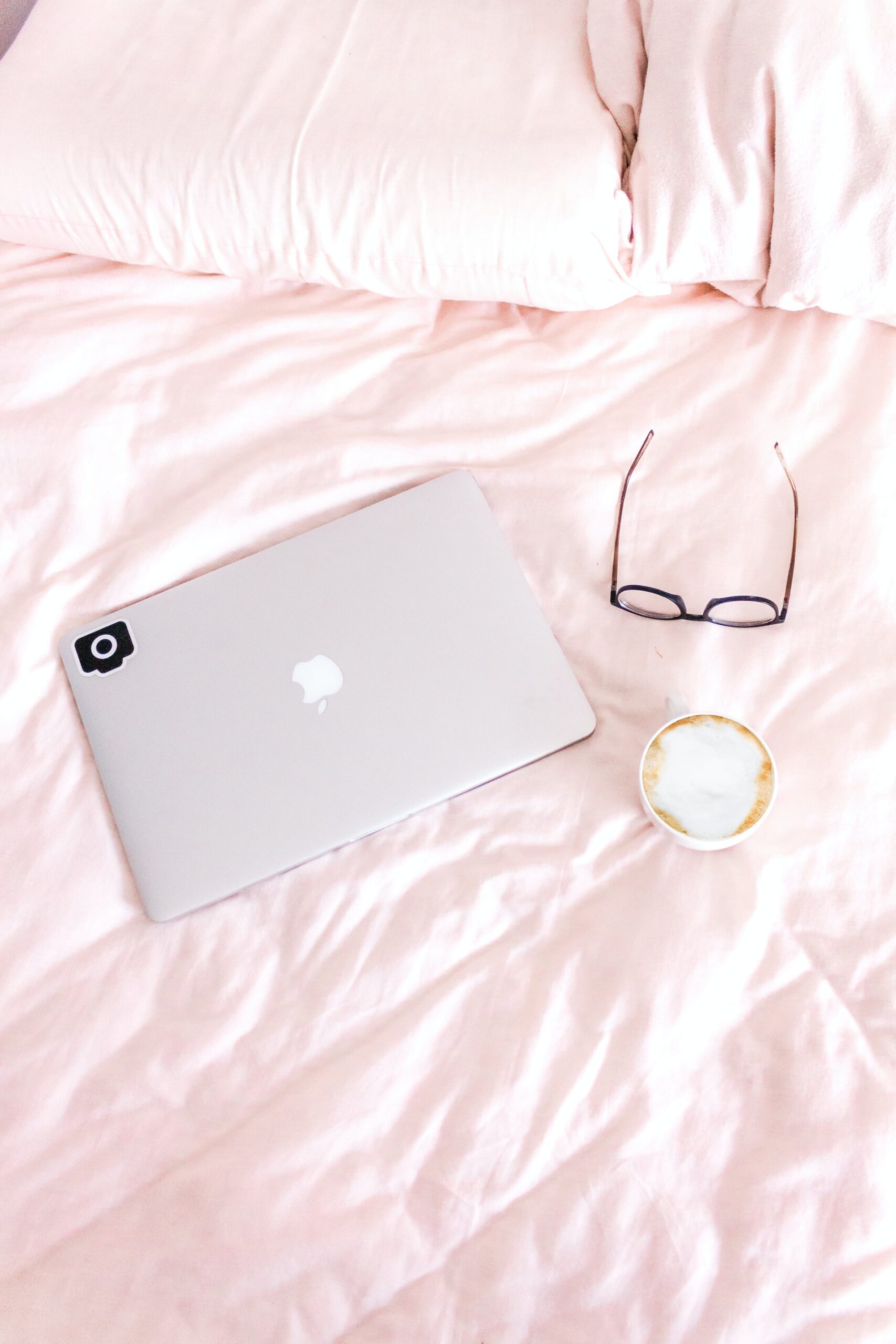 A photographer's laptop laying on a pink bedspread with coffee and a pair of glasses